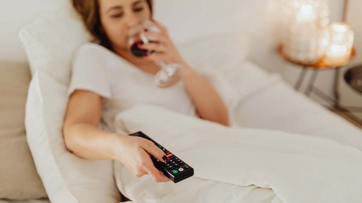 A woman in bed holding a glass of wine and a remote control, enjoying a cozy evening.